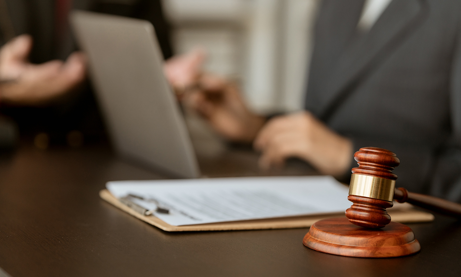 a gavel in the foreground with paperwork and two people sitting around a laptop in the background