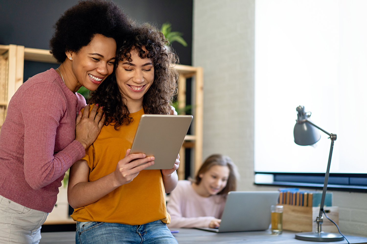 two women smiling, looking at a tablet, and a woman smiling at the computer on a table